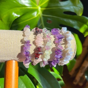 K&ouml;nnte beinhalten: F&uuml;nf Edelstein-Armb&auml;nder, die auf einem Holzst&auml;nder gestapelt sind. Die Armb&auml;nder bestehen aus Amethyst, Rosenquarz, gr&uuml;nem Aventurin, Granat und blauer Spitze.