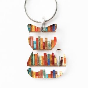 May include: A silver key chain with a cat-shaped charm. The cat is made of clear acrylic and has a bookshelf design with colorful books.