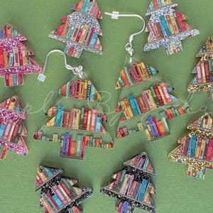 May include: A collection of Christmas tree-shaped earrings and charms. The trees are decorated with colorful book spines and glitter. The earrings have silver hooks. The background is a solid green.