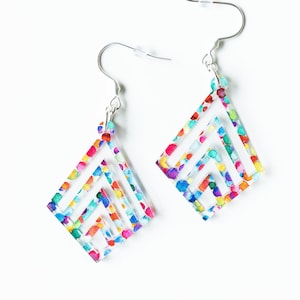 May include: A pair of colorful geometric earrings. Each earring features a clear, diamond-shaped design with a layered, open structure. The earrings are adorned with a vibrant pattern of multicolored dots, and are suspended from silver-colored hooks.