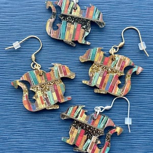 May include: A pair of gold and silver earrings featuring a dragon design with a bookshelf pattern. The dragon is made of wood and has a glittery finish.
