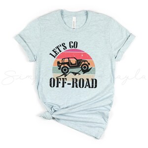 May include: A light blue t-shirt with the text "Let's Go Off-Road" in black. A black silhouette of a jeep driving over a mountain range is in a sunset-colored circle behind the text.