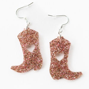 May include: A pair of pink glitter cowboy boot earrings with a heart cut-out. The earrings have silver hooks.