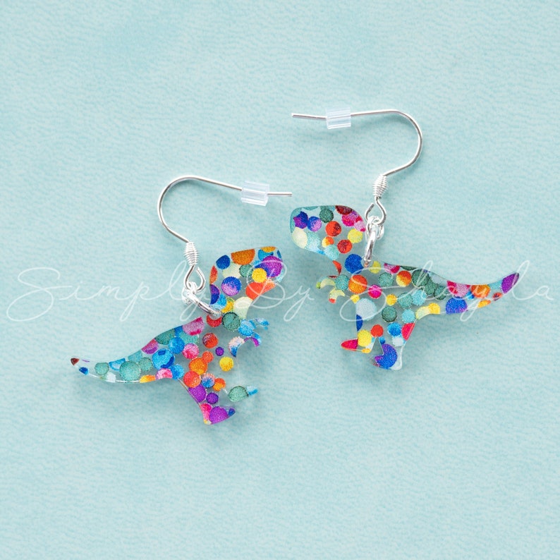 May include: Dinosaur-shaped earrings with a colorful confetti pattern. The earrings are made of a clear material with a silver hook. The dinosaur design features a variety of colors, including blue, orange, yellow, and purple.