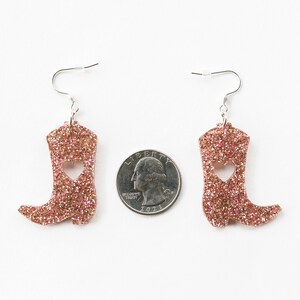 May include: A pair of pink glitter cowboy boot earrings with a heart cut-out. The earrings are approximately the size of a quarter.