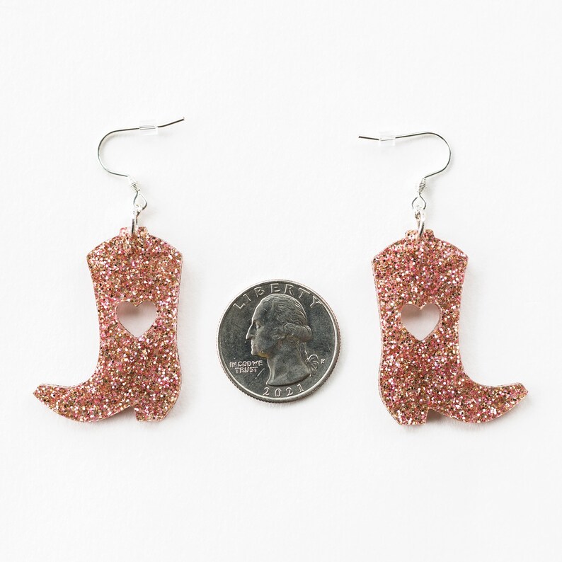 May include: A pair of pink glitter cowboy boot earrings with a heart cut-out. The earrings are approximately the size of a quarter.