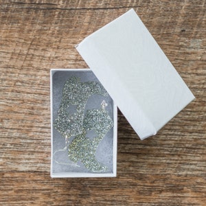 May include: A pair of silver glitter boot earrings in a white gift box. The earrings are shaped like boots with a star on each boot.