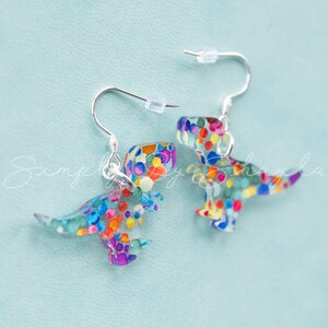 May include: A pair of dinosaur-shaped earrings with silver hooks. The earrings are clear with colorful confetti-like accents. The dinosaur shapes are facing opposite directions, with one slightly out of focus.