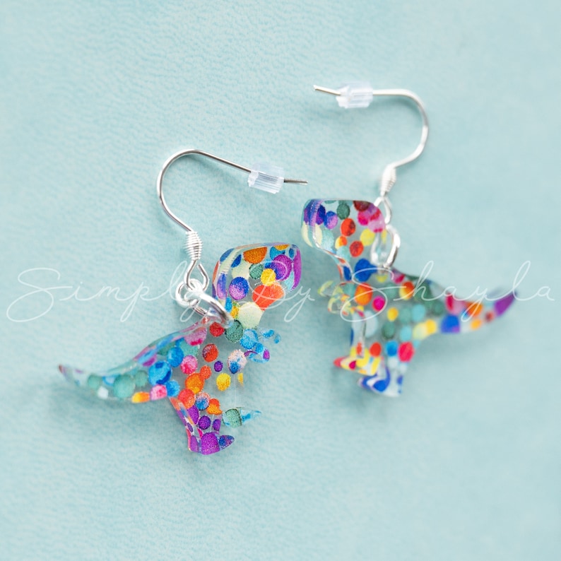 May include: A pair of dinosaur-shaped earrings with silver hooks. The earrings are clear with colorful confetti-like accents. The dinosaur shapes are facing opposite directions, with one slightly out of focus.