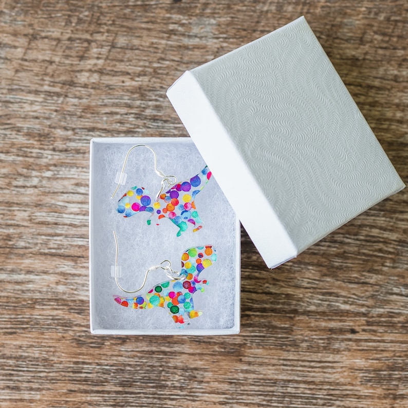 May include: A pair of dinosaur-shaped earrings with colorful polka dots, presented in a white gift box. The earrings have silver-colored hooks. The box is open, revealing the earrings nestled on white filler, set against a wooden surface.