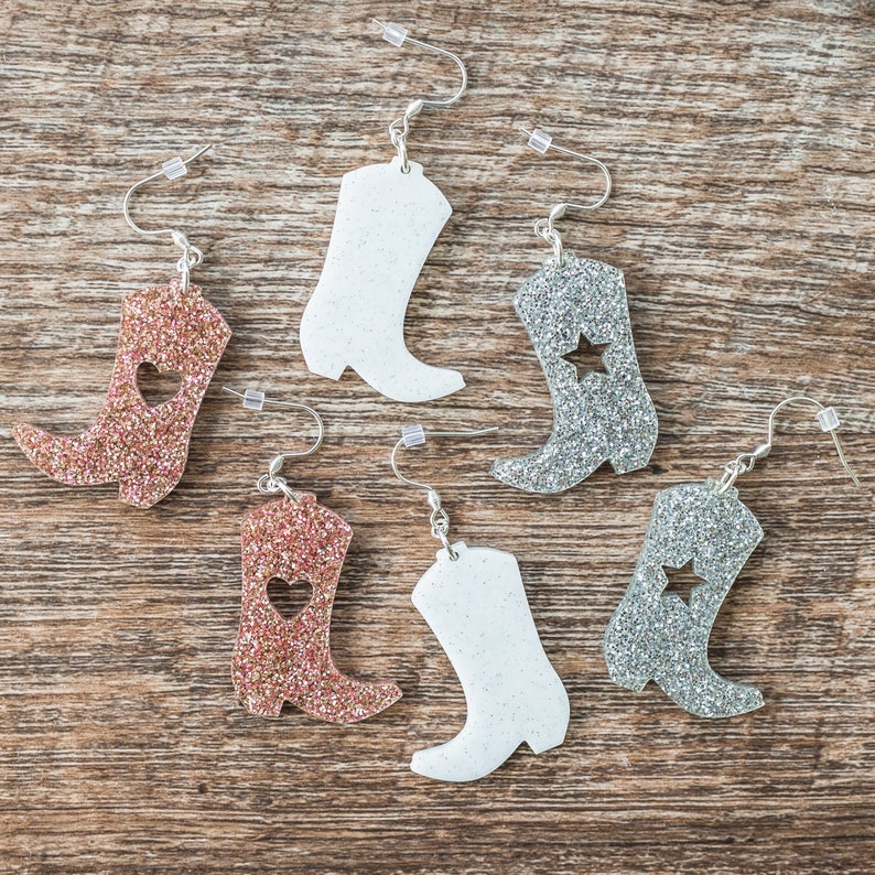 May include: Six pairs of glitter boot earrings. Two pairs are silver glitter with a star cutout, two pairs are rose gold glitter with a heart cutout, and two pairs are white glitter.