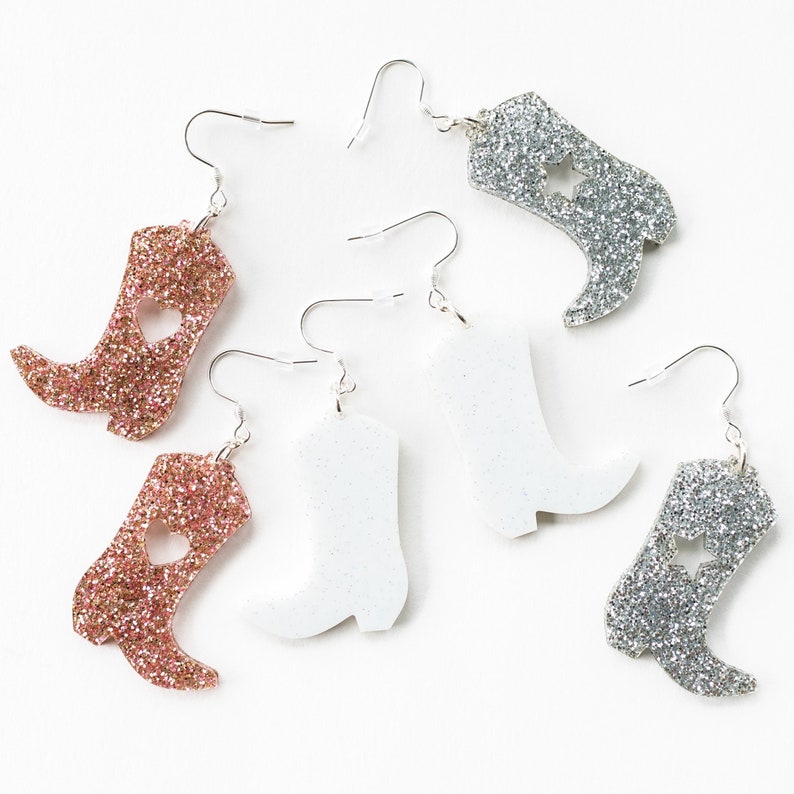 May include: Five pairs of dangle earrings featuring a boot design. Two pairs are silver glitter, one pair is white glitter, and two pairs are pink glitter. Each boot has a different design, including a heart and a star.