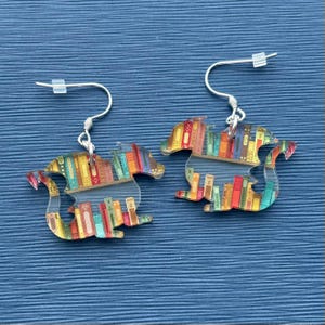 May include: A pair of silver dangle earrings featuring a dragon silhouette with a bookshelf and colorful books inside. The dragon is facing to the right.
