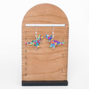 May include: A pair of dinosaur-shaped earrings with colorful polka dots. The earrings are made of a clear material with a silver hook. The earrings are displayed on a wooden stand with a ruler.