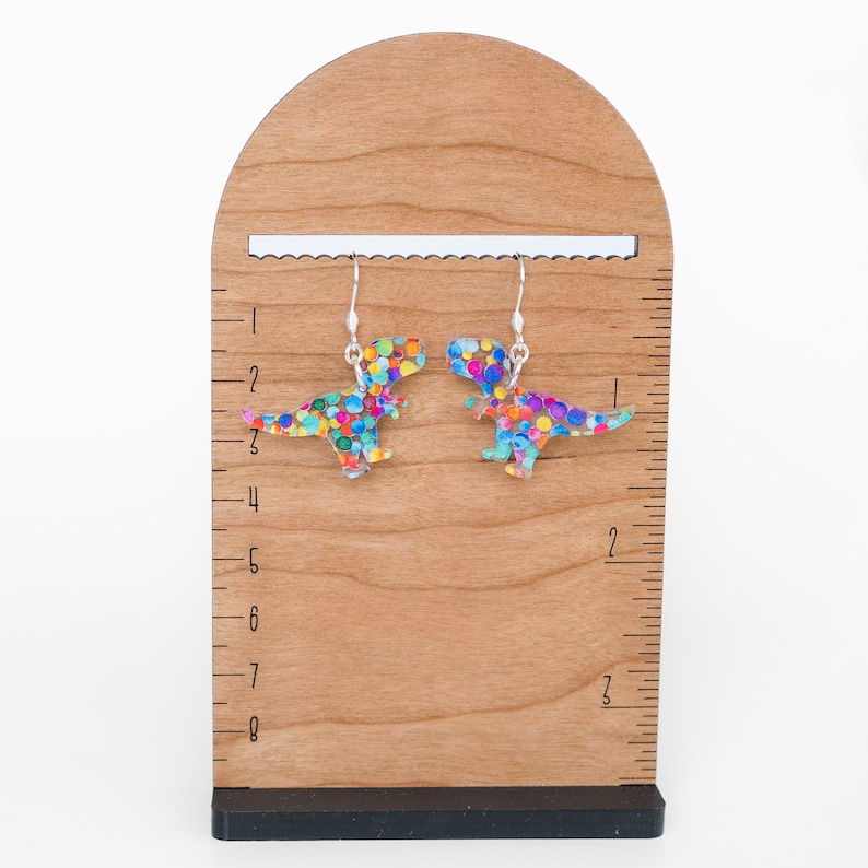 May include: A pair of dinosaur-shaped earrings with colorful polka dots. The earrings are made of a clear material with a silver hook. The earrings are displayed on a wooden stand with a ruler.