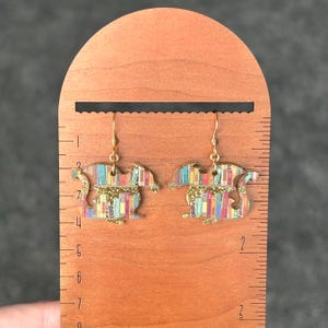 May include: A pair of gold-toned earrings featuring a dragon silhouette with a colorful book-themed design. The earrings are shown against a wooden ruler.