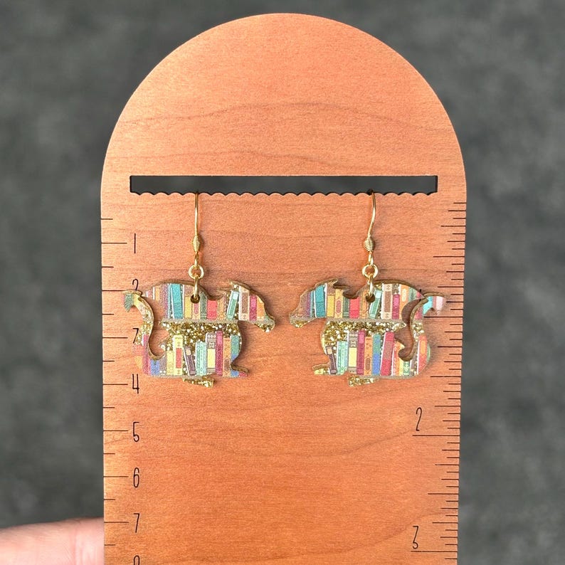 May include: A pair of gold-toned earrings featuring a dragon silhouette with a colorful book-themed design. The earrings are shown against a wooden ruler.