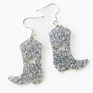 May include: Silver glitter cowboy boot earrings with a star cut-out. The earrings have silver hooks.