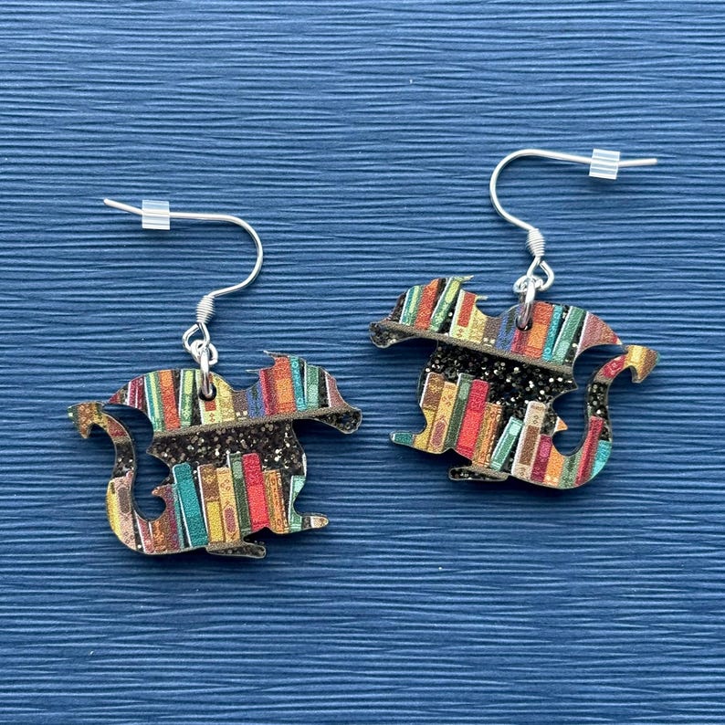 May include: A pair of silver dangle earrings featuring a dragon silhouette with a bookshelf design. The bookshelf is filled with colorful books and has a glittery black background.