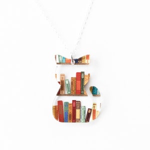 May include: A clear cat-shaped pendant necklace with a silver chain. The pendant is filled with colorful books, and the cat's head and tail are decorated with a patterned design. The necklace is on a white background.