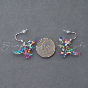 May include: A pair of dinosaur-shaped earrings with colorful confetti-like patterns. The earrings have silver-colored hooks. The image is on a gray background.