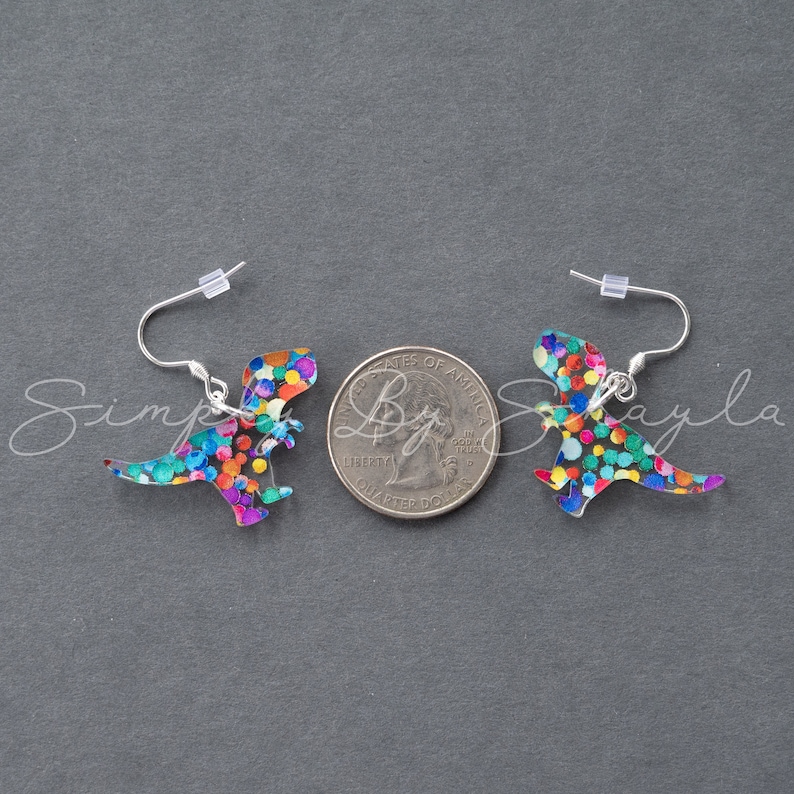 May include: A pair of dinosaur-shaped earrings with colorful confetti-like patterns. The earrings have silver-colored hooks. The image is on a gray background.
