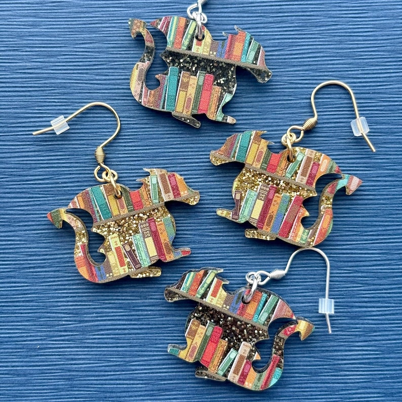 May include: A pair of gold and silver earrings featuring a dragon design with a bookshelf pattern. The dragon is made of wood and has a glittery finish.