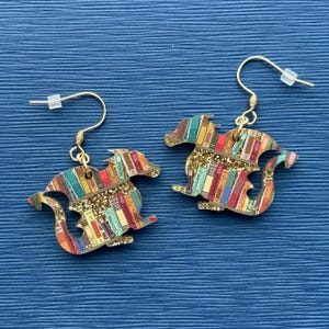 May include: A pair of gold dangle earrings featuring a dragon silhouette made of wood. The dragon is covered in a colorful pattern of books and gold glitter.