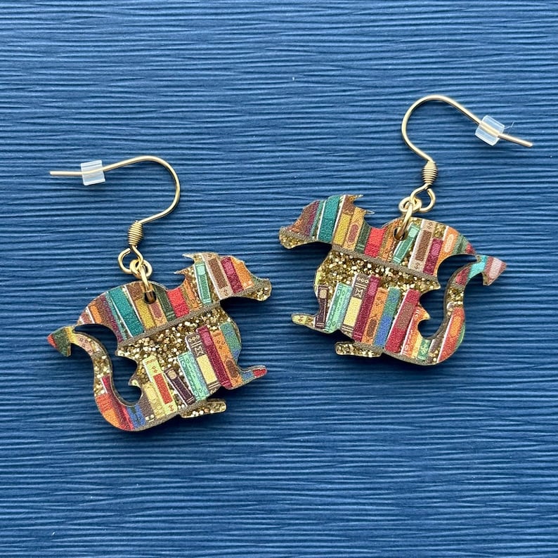 May include: A pair of gold dangle earrings featuring a dragon silhouette made of wood. The dragon is covered in a colorful pattern of books and gold glitter.
