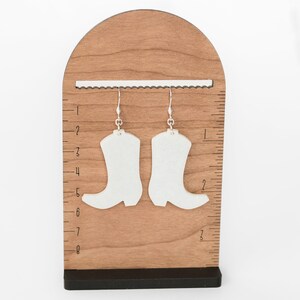 May include: A pair of white boot-shaped earrings with silver hooks hanging from a wooden display stand with a ruler.
