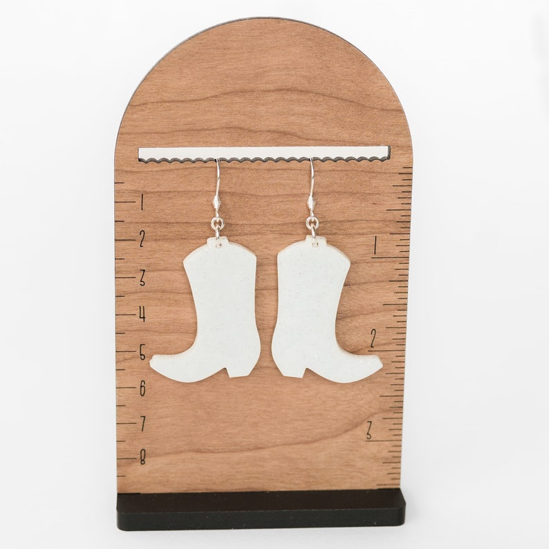 May include: A pair of white boot-shaped earrings with silver hooks hanging from a wooden display stand with a ruler.