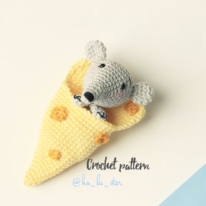 Op de afbeelding: Een grijze gehaakte muis slaapt in een gele gehaakte kaaswig. De kaaswig heeft een bruin steekdetail. De tekst "Crochet pattern @ha_lu_ster" staat onder de muis.