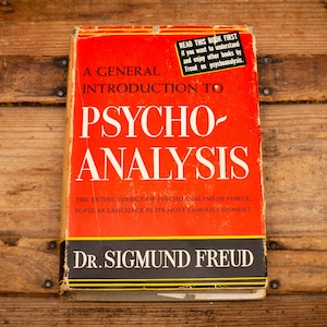 May include: A vintage book titled "A General Introduction to Psychoanalysis" by Dr. Sigmund Freud. The book has a red cover with white lettering and a black and yellow border. A sticker reads "Read this book first".