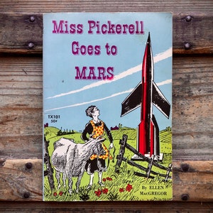 May include: Vintage book cover for "Miss Pickerell Goes to Mars" featuring a retro illustration. The cover shows a woman in an apron, a cow, a rocket, and the title text. The book is set against a blue sky background.