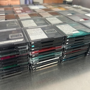 May include: A large stack of square Maxell MD 74 MiniDiscs in black plastic cases. Several discs feature white adhesive labels with handwritten black ink text, including dates and song titles. The cases are stacked in multiple rows and columns.