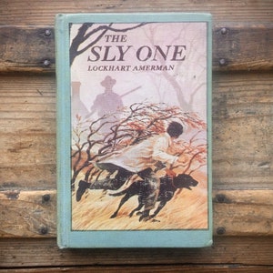 May include: Vintage book titled "The Sly One" by Lockhart Amerman. The cover features an illustration of a man running with a dog through a field, with a hunter in the background. The book has a light blue spine and is set against a wooden background.