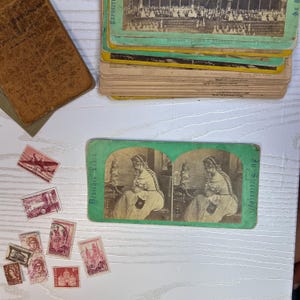 Puede incluir: Una colección de objetos antiguos, que incluye tarjetas estereoscópicas, sellos y un libro encuadernado en cuero. Las tarjetas estereoscópicas presentan imágenes de una mujer y escenas de una exposición. Los sellos muestran varios diseños y colores.