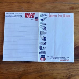 Può includere: Un blocco note bianco con linee orizzontali blu e le parole "LOAD 'EM UP... SHIP 'EM OUT" e "PIGGY BACK" stampate in nero e rosso. Il lato destro presenta illustrazioni di vagoni ferroviari e il testo "EQUIPPED FOR SERVICE". È visibile anche il logo Union Pacific.