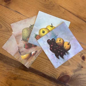 May include: A set of square coasters with fruit illustrations. The coasters depict pears, apples, strawberries, and cherries. The color scheme includes green, red, yellow, and brown hues. The coasters are displayed on a wooden surface.