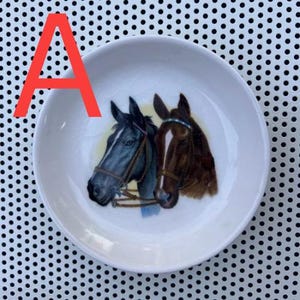 May include: A white ceramic saucer with a black and brown horse design. The horses are facing each other and have bridles on.