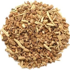 May include: A pile of dried, chopped botanical root pieces in various shades of light and medium brown. The texture is woody, fibrous, and irregular, consisting of small, fragmented bits and thin, splinter-like strips arranged in a circular mound.
