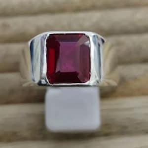 May include: A silver ring with a large, rectangular, deep red gemstone. The gemstone is set in a polished silver band, creating a classic design. The ring is displayed on a white surface against a neutral background.