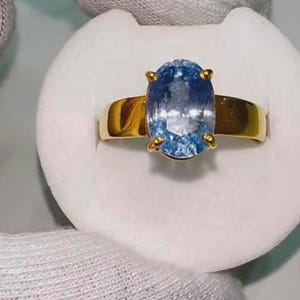 Könnte beinhalten: Ein goldener Ring mit einem großen, oval geschliffenen, hellblauen Edelstein. Der Edelstein wird von vier Krappen gehalten. Der Ring ist aus schlichtem, poliertem Gold. Der Ring wird auf einer weißen Oberfläche präsentiert.