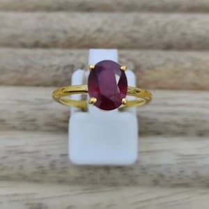 May include: A gold ring with a large oval-shaped ruby gemstone. The ring is set on a white display stand.