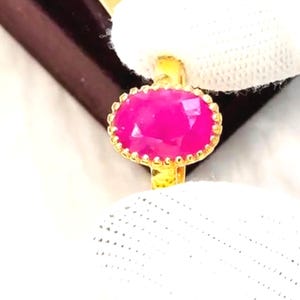 Könnte beinhalten: Ein goldener Ring mit einem großen, ovalen, leuchtend rosa Edelstein. Der Edelstein ist von einer goldenen Lünette mit kleinen dekorativen Details umgeben. Der Ring ist vor einem weißen Hintergrund platziert.