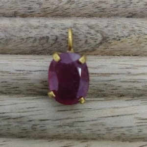 May include: A gold pendant with a deep red oval gemstone set in a prong setting.
