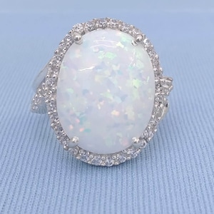 Puede incluir: Un anillo de plata con una gran piedra ovalada de ópalo blanco rodeada por un halo de piedras más pequeñas y transparentes.