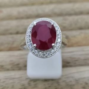 May include: A silver ring with a large oval ruby gemstone surrounded by a halo of smaller white gemstones.