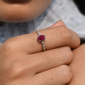 Può includere: Un anello d'argento con un grande rubino ovale come pietra centrale. La fascia è impreziosita da pietre preziose più piccole e trasparenti. L'anello è mostrato su un dito, evidenziando il suo design e il suo colore.