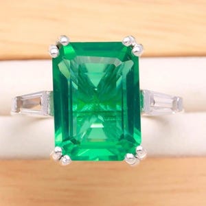 Könnte beinhalten: Ein silberfarbener Ring mit einem großen, rechteckigen, smaragdgeschliffenen grünen Edelstein, der von vier Krappen gehalten wird. Das Band ist auf jeder Seite mit einem kleineren, klaren, im Baguetteschliff geschliffenen Stein besetzt, was ein klassisches Drei-Stein-Design ergibt.